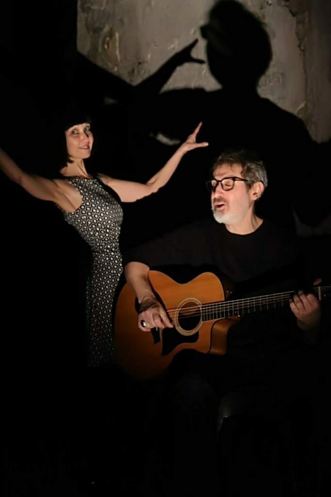 Une femme danse et un homme joue de la guitare, éclairé de face par une faible lumière. La femme sourit et semble heureuse.