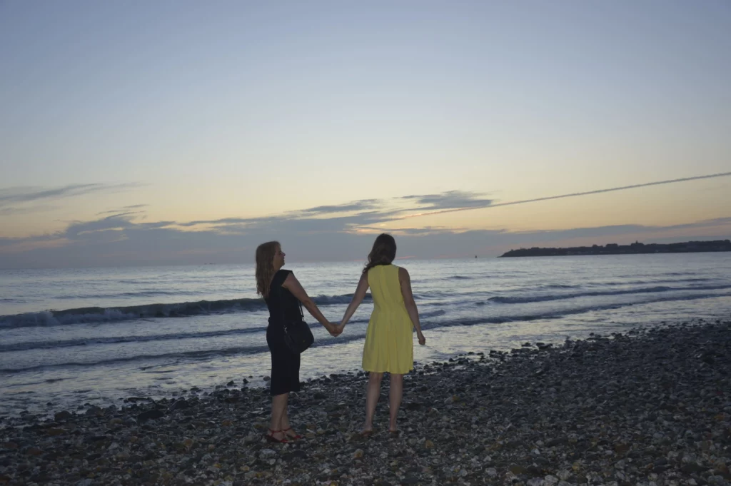 Deux femmes, l'une à la robe noir, l'autre à la robe jaune, se tiennent la main sur une plage de galets. Le soleil semble se coucher en arrière plan, au dessus de la mer.