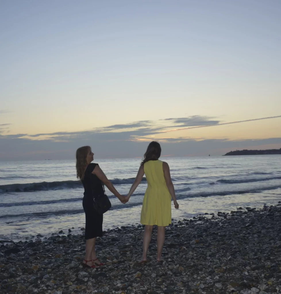 Deux femmes, l'une à la robe noir, l'autre à la robe jaune, se tiennent la main sur une plage de galets. Le soleil semble se coucher en arrière plan, au dessus de la mer.