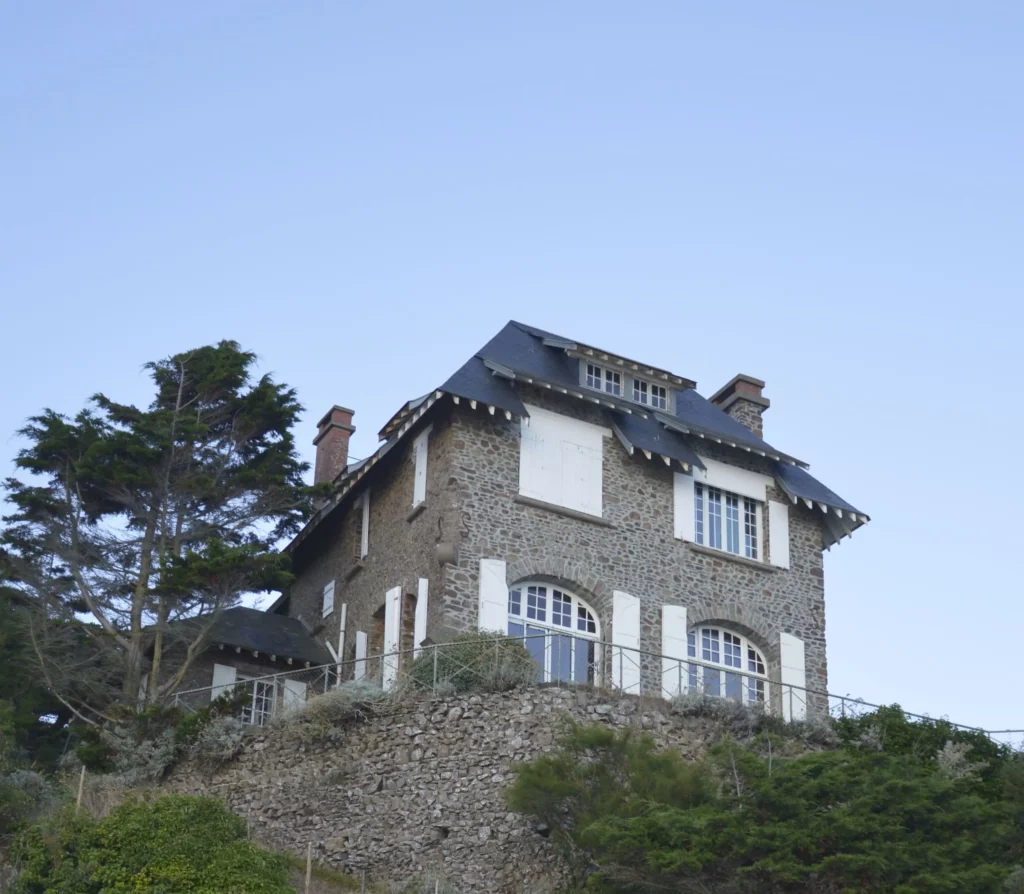 Une maison de pierre, en haut d'une falaise, un arbre à sa gauche et le ciel bleu en arrière-plan.